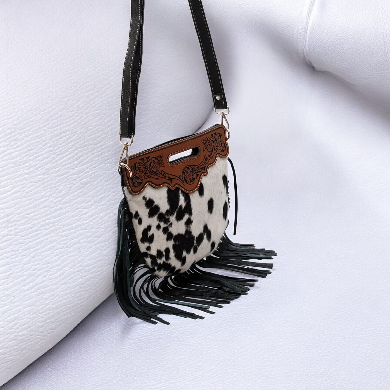 Cow print handbag with black fringe and brown leather accents on a white background