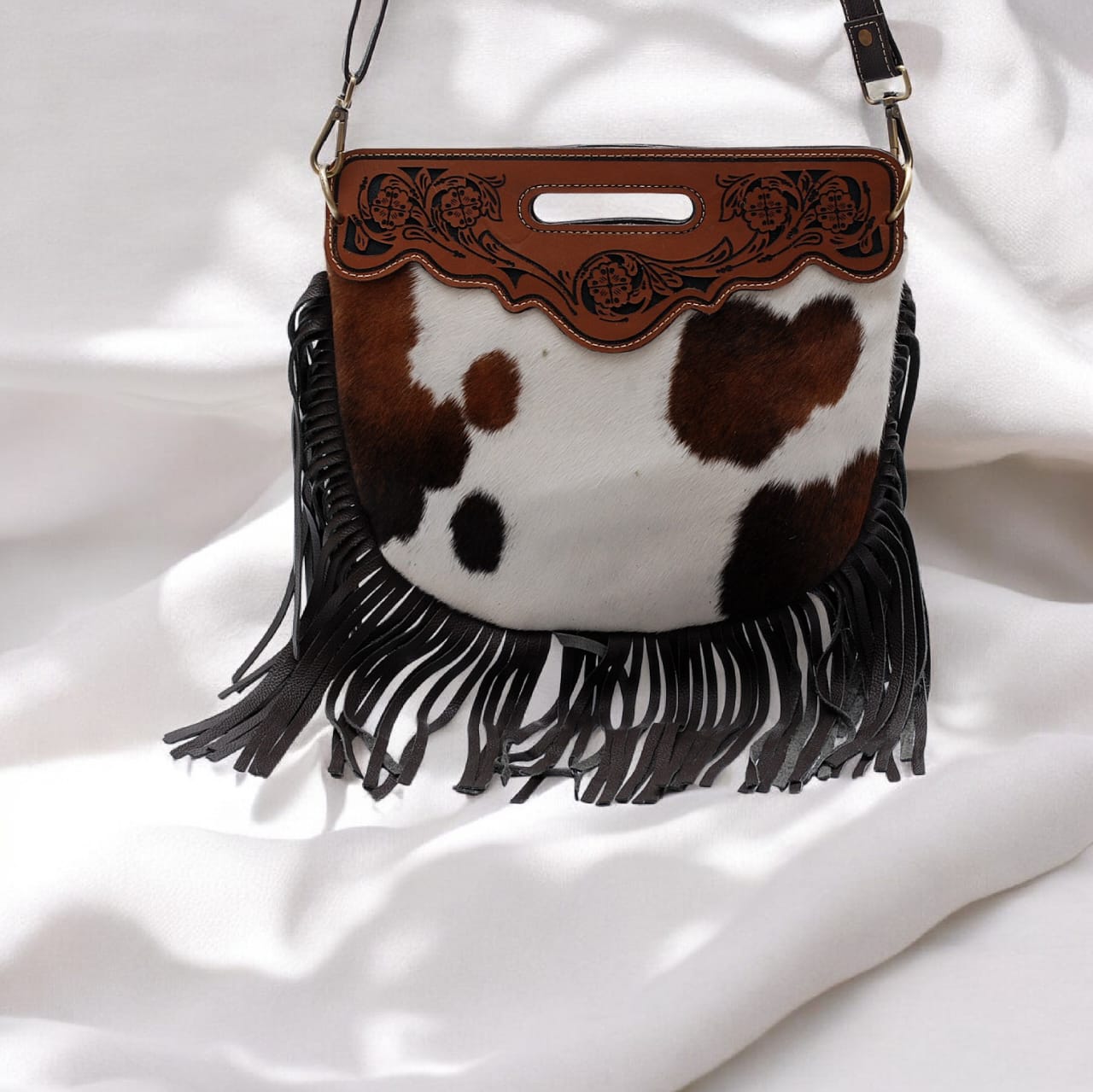 Cow print handbag with brown leather top and black fringe on a white background
